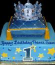 Prince 1st Birthday Cake,  Blue buttercream iced,  square decorated with stars, dots, and a tasseled pillow with white pearls. A jeweled crown tops this royal cake off.  Everything on this cake is EDIBLE. (Serves 12-90 party slices)